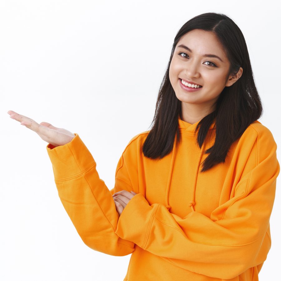 Waist-up portrait cute beautiful asian woman in orange hoodie, introduce produce banner, pointing at blank white space, holding product and smiling, recommend advertisement, white background.